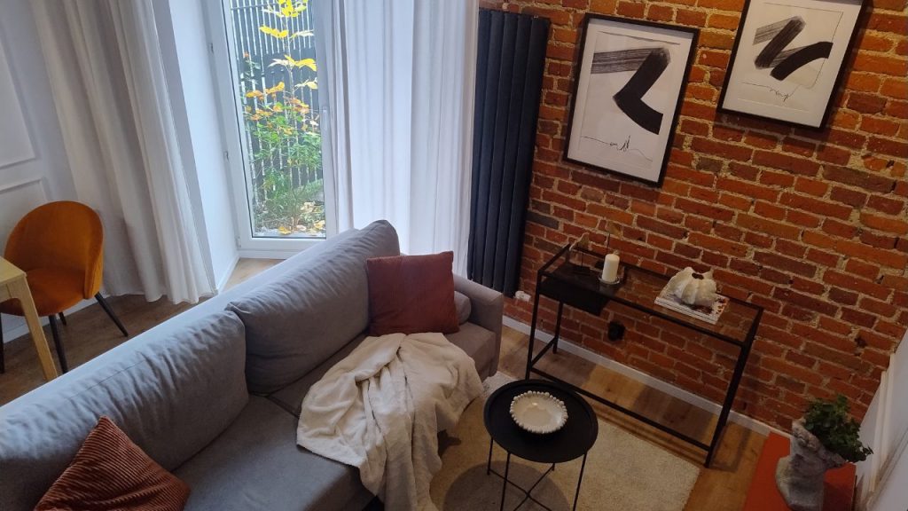 Wnętrze mieszkania w bieli i czerni ocieplone ścianą z cegły, na niej plakaty ze sztuka abstrakcyjną, szara sofa z rudymi poduszkami