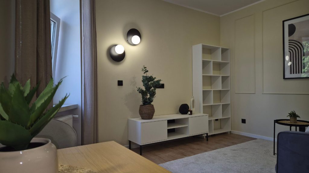 Wnętrze kawalerki w stonowanych , jasnych barwach, sztukateria, dekoracyjny plakat, designerskie oświetlenie, aranżacja ściany tv z szafką i regałem